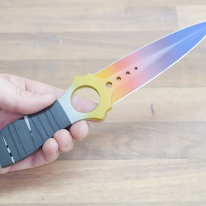 May include: A rainbow-colored knife with a black handle and a ring on the handle. The blade is a gradient of yellow, orange, red, and blue.