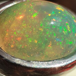 May include: A close-up of a green opal gemstone set in a silver ring. The opal has a bright, iridescent sheen with flashes of red, orange, and yellow.