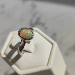 May include: A silver ring with a light green opal gemstone. The ring is on a white display stand.