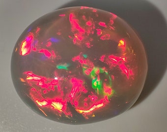 May include: A polished, round opal gemstone with a dark base color. The opal displays vibrant flashes of red, pink, green, and yellow throughout its surface. The gemstone is smooth and reflective, with a subtle shadow.