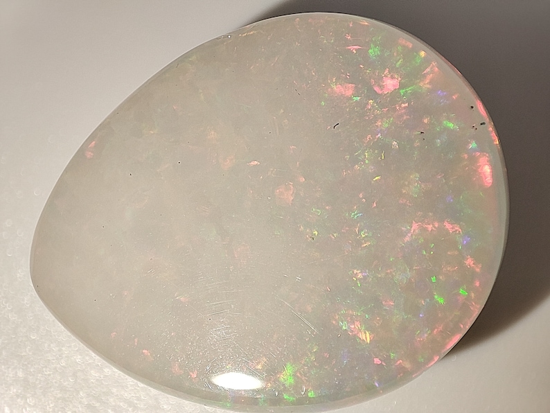 May include: A white opal gemstone with a teardrop shape and iridescent flashes of pink, green, and blue.