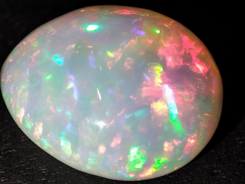May include: A white opal gemstone with a smooth, rounded shape. The opal displays a variety of iridescent colors, including green, blue, pink, and yellow.