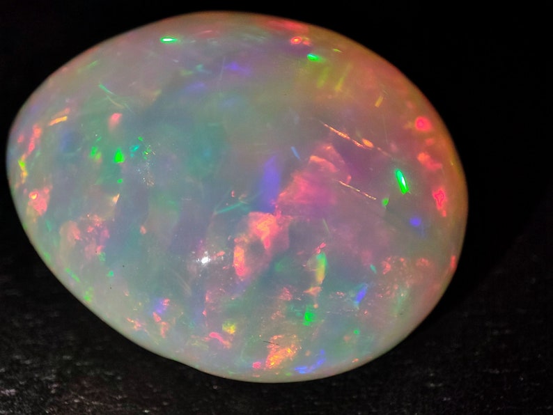 May include: A white opal gemstone with a smooth, oval shape. The opal displays a variety of iridescent colors, including green, blue, red, and orange.