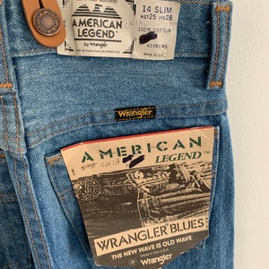 May include: A pair of blue denim jeans with a brown leather belt loop and a Wrangler American Legend tag. The tag has the text "Wrangler Blues" and "The New Wave is Old Wave".