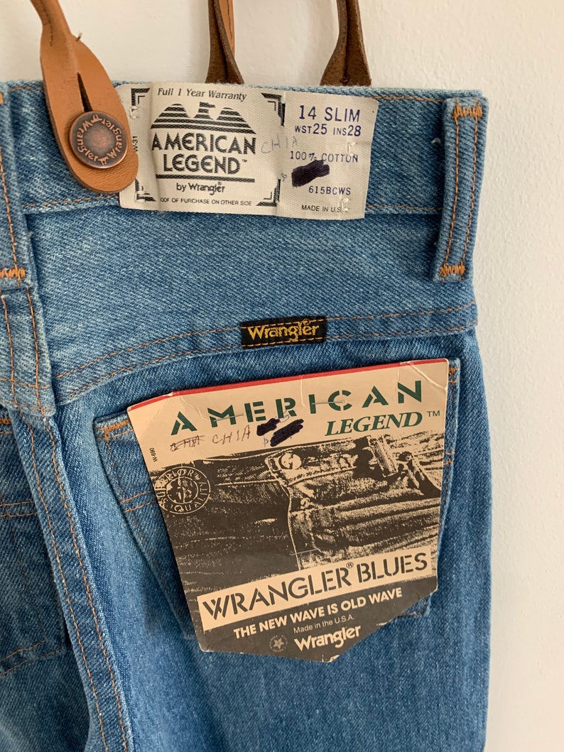 May include: A pair of blue denim jeans with a brown leather belt loop and a Wrangler American Legend tag. The tag has the text "Wrangler Blues" and "The New Wave is Old Wave".