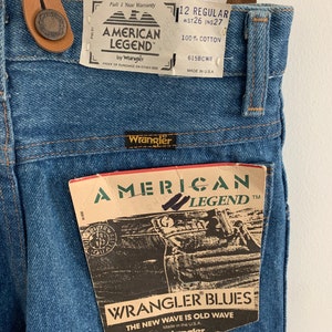 May include: Blue denim dungarees with a brown leather strap and buckle. The dungarees have a white label that says "American Legend by Wrangler" and a smaller label that says "12 Regular WST26 INS27 100% Cotton 615BCWR Made in USA".