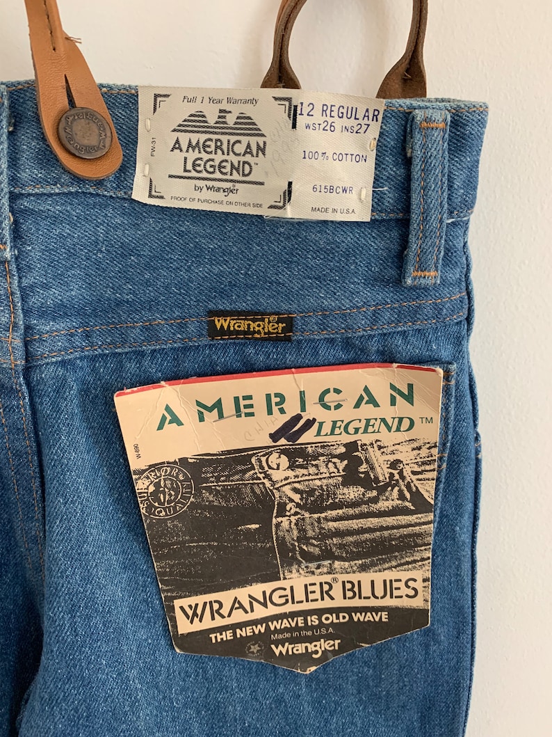 May include: Blue denim dungarees with a brown leather strap and buckle. The dungarees have a white label that says "American Legend by Wrangler" and a smaller label that says "12 Regular WST26 INS27 100% Cotton 615BCWR Made in USA".
