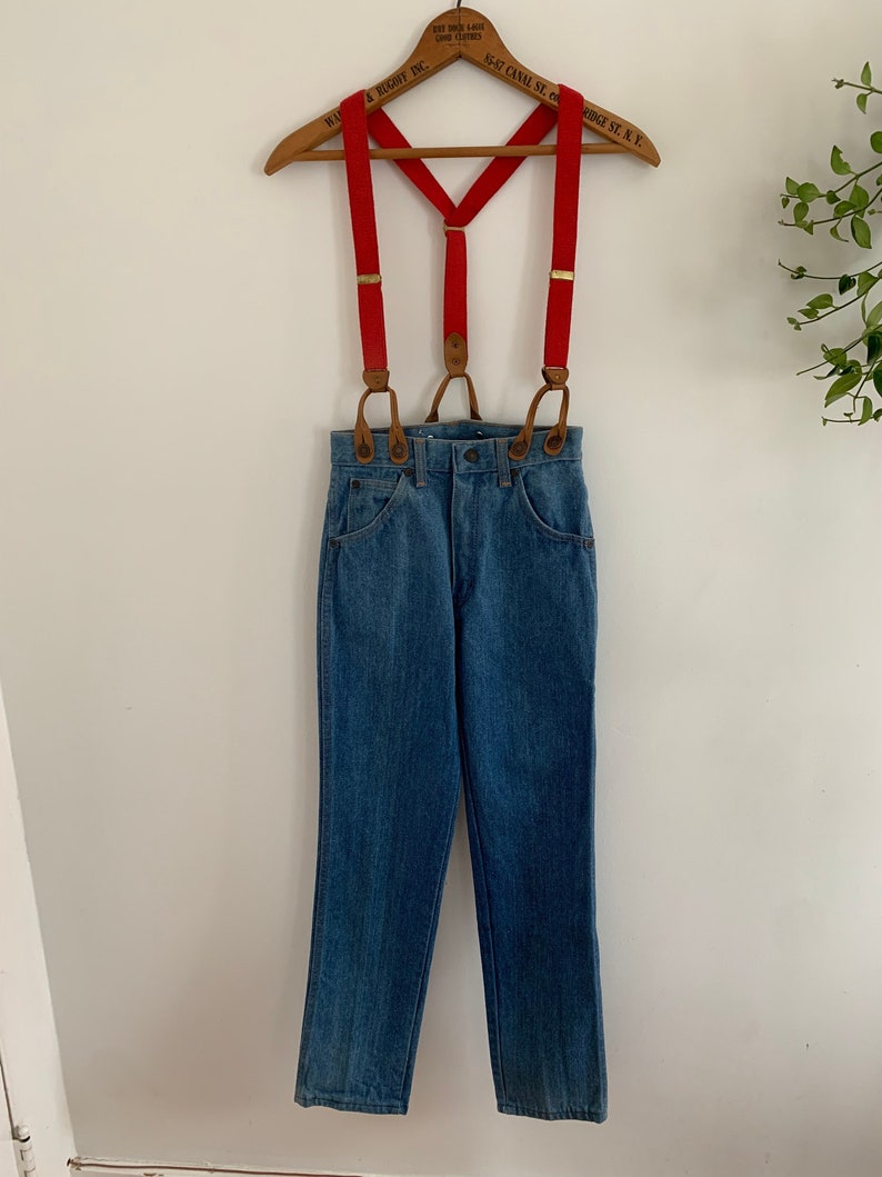 May include: Red braces with brown leather accents hanging on a wooden hanger with blue denim jeans.