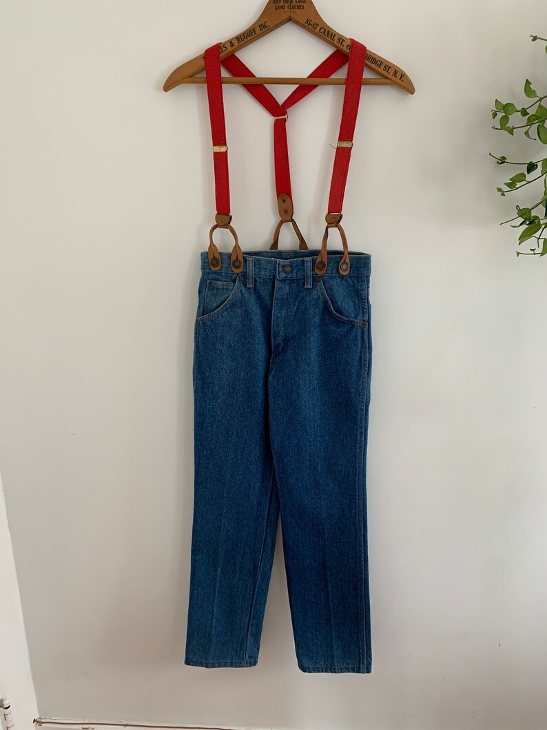 May include: Red braces with brown leather accents and a pair of blue denim jeans.