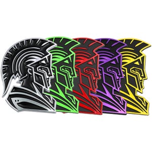 Puede incluir: Cinco emblemas de casco espartano de metal en diferentes colores: negro, verde, rojo, morado y amarillo. Cada casco está mirando hacia la derecha y tiene un diseño detallado.