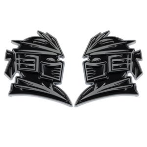 Puede incluir: Dos calcomanías de emblema negras y plateadas con un diseño de casco de guerrero estilizado. Las calcomanías tienen un acabado brillante sobre un fondo blanco. El diseño incluye ángulos agudos y formas geométricas.