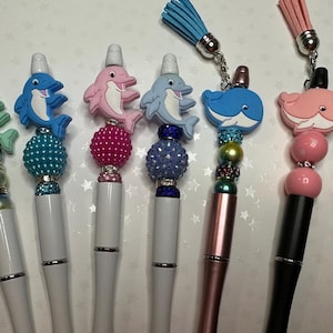 May include: A collection of decorative pens featuring dolphin and whale toppers in various colors. The pens have white or metallic barrels, embellished with beads and tassels. The toppers include green, blue, and pink dolphins, and blue and pink whales.