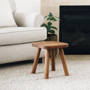 May include: A small, square wooden stool with four rounded legs. The stool is made of light brown wood and sits on a light-colored carpet. The background includes a neutral-toned armchair, a fireplace, and a potted plant.