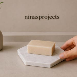 May include: A rectangular beige bar of soap rests on a white hexagonal marble tray. To the left, a small beige ceramic vase holds a green leafy sprig. The text ninasprojects is visible in the background. The composition is neutral and clean.
