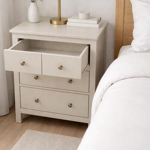 May include: A light beige nightstand with three drawers, one open, featuring brass knobs. A white lamp with a gold base sits on top, along with books and a vase with greenery. A white bed and rug are in the frame.