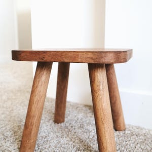 May include: A small, round wooden stool with three angled legs. The stool is made of a light brown wood and has a simple, minimalist design. The legs are sturdy and slightly tapered, providing stability. The stool is suitable for various uses.