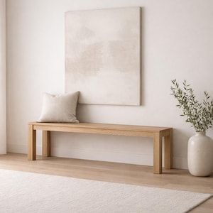 May include: A light-colored wooden bench with a square cushion, set against a white wall. A large, neutral-toned abstract painting hangs above the bench. A white vase with greenery sits to the right, and a light-colored rug is on the floor.