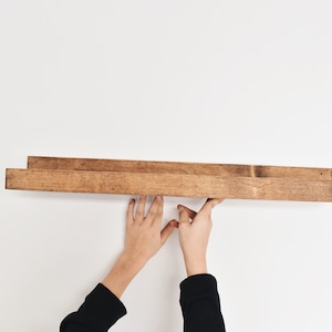 May include: A person is installing a wooden shelf on a white wall. The shelf is made of two pieces of wood, each about 1.5 inches thick. The shelf is stained a dark brown color.