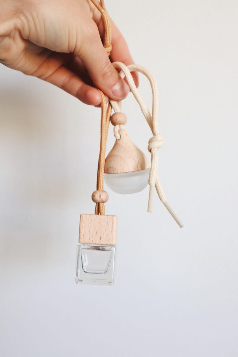 Puede incluir: Un ambientador de coche de madera con una botella de vidrio transparente y una tapa de madera. El ambientador est&aacute; colgado de una cuerda beige y marr&oacute;n.