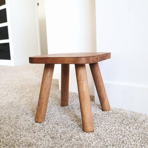 May include: A small wooden stool with four legs. The stool is made of dark brown wood and has a square top.