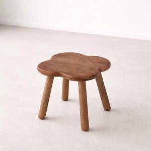 May include: A small, wooden stool with a four-leaf clover shaped seat. The stool has four rounded legs and is made of a warm-toned wood. The design is simple and minimalist, suitable for various interior styles.