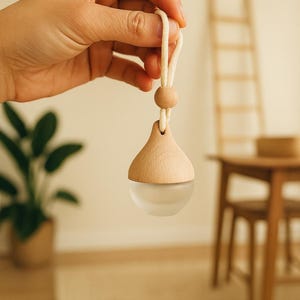 May include: A wooden essential oil diffuser with a frosted glass base, suspended by a cream-colored cord and wooden bead. The teardrop-shaped diffuser is held up by a hand. The background is blurred, with a plant and wooden furniture visible.