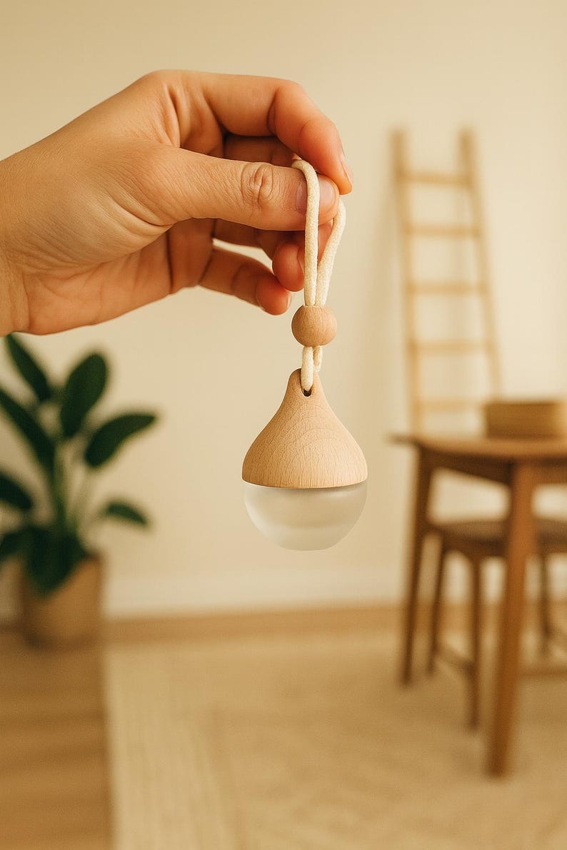 K&ouml;nnte beinhalten: Ein &auml;therisches &Ouml;l-Diffusor aus Holz mit einer Basis aus mattiertem Glas, aufgeh&auml;ngt an einer cremefarbenen Schnur und einer Holzperle. Der tropfenf&ouml;rmige Diffusor wird von einer Hand gehalten. Der Hintergrund ist verschwommen, mit einer Pflanze und Holzm&ouml;beln.