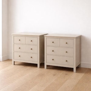 May include: Two matching beige dressers with four drawers each, featuring brass knobs. The dressers are set against a white wall and sit on a light-colored wooden floor. The design is simple and classic.