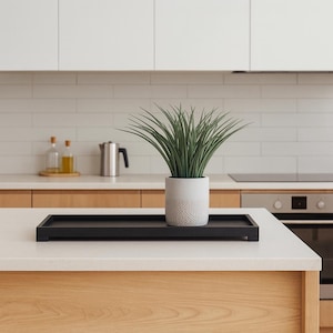 May include: A black rectangular tray with a potted green plant on a white countertop. The plant is in a white textured pot. The kitchen has white cabinets and a light wood base.