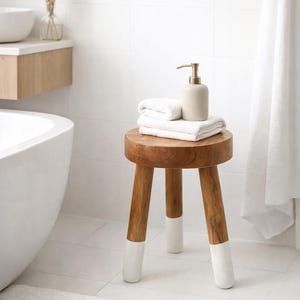 May include: A wooden stool with three legs, each tipped with white paint, holds a stack of white towels and a beige soap dispenser with a gold pump. The stool is in a white-tiled bathroom, next to a white bathtub.