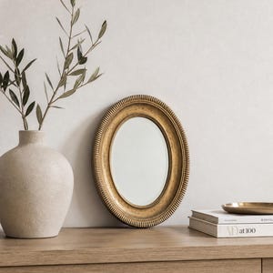 May include: An oval, gold-framed mirror with a textured border sits on a wooden surface. A beige vase holds leafy branches to the left. A stack of books and a small gold tray are also on the surface, creating a minimalist aesthetic.