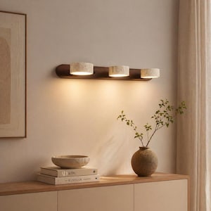 May include: A modern wall-mounted light fixture with three cylindrical lamps. The lamps are made of a speckled, light-colored material and are mounted on a dark brown wooden base. The fixture is installed on a neutral-colored wall, with a warm glow.