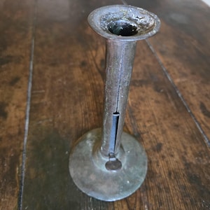 May include: A vintage metal candlestick holder with a wide, circular base and a tall, cylindrical stem. The top flares out into a shallow cup. The metal has a tarnished, aged appearance, with visible wear and discoloration, suggesting its antique nature.