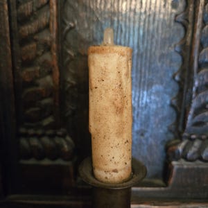 May include: A cylindrical, off-white candle with a textured, weathered surface featuring dark brown speckles and drips. The candle is set into a dark metal holder, displaying a worn, aged appearance with a small, translucent wick at the top.