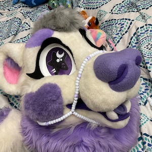 May include: A white and purple lion fursuit head with a star and moon design in the eyes. The fursuit has a purple nose, ear accents, and a beaded necklace. The fur is white and purple.