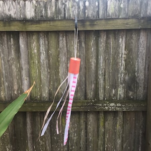 May include: A red and white striped windsock hanging from a hook on a wooden fence. The windsock is made from a can with ribbons attached.