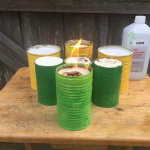 May include: Six painted metal cans with flames burning inside. The cans are painted yellow and green. A bottle of 70% isopropyl alcohol is on the table.