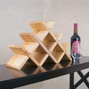 May include: A light-colored wooden wine rack with a geometric, interlocking design featuring multiple triangular storage compartments. Beside the rack stands a dark glass wine bottle with a red label and a clear glass tumbler.