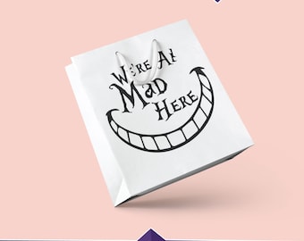 Può includere: Una borsa di carta bianca con un ghigno nero di Gatto del Cheshire e il testo "We're All Mad Here".