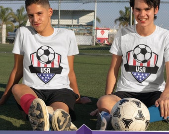 May include: Two people wearing white t-shirts with a soccer ball and USA flag design. The shirts have the word "USA" printed on them. The people are sitting on a green grassy field with a soccer ball in front of them.