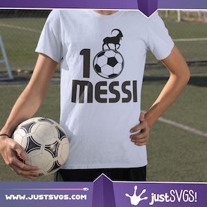 May include: A white t-shirt with a black graphic of a soccer ball with a goat standing on top of it. The number 1 is to the left of the ball and the word "MESSI" is below the ball.