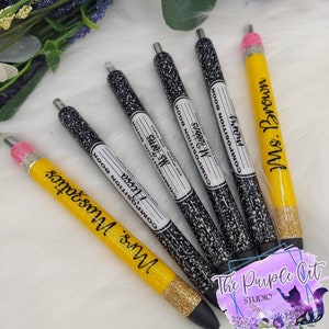 May include: A collection of pens with a composition book design and yellow pencil-shaped pens. The composition book pens have black and white speckled barrels with a label that reads "Composition Book" and teacher names. The yellow pens have glitter and teacher names.