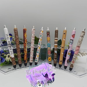 May include: A collection of ten pens with various coffee-themed designs. The pens feature patterns like coffee beans, donuts, and coffee cups. One pen has the text "but coffee first...". The pens are displayed in a clear acrylic holder.