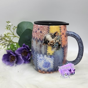 May include: A glittery tumbler with a black lid and handle, featuring a patchwork design of pink, yellow, and blue panels. The surface displays a cartoon lamb holding flowers, small paw prints, and floral patterns. A logo reads The Purple Cat Studio.