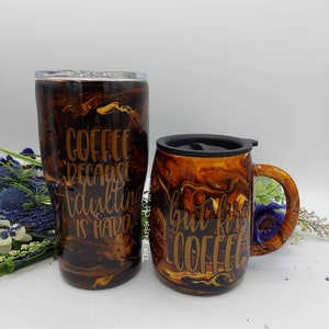 May include: Two coffee-themed tumblers and mugs with a brown and gold marbled design. One tumbler reads "Coffee because adulting is hard." The other mug reads "but first coffee."