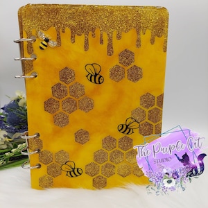 Puede incluir: Un diario hecho a mano con un tema de abejas y panales. El diario presenta una combinación de colores amarillo y dorado, con una parte superior dorada brillante que se asemeja a la miel que gotea. Ilustraciones de abejas negras y patrones de panal decoran el frente.