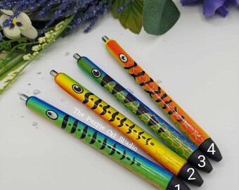 May include: Four colorful pens with fish-scale designs. Each pen has a different color scheme, including blue, green, yellow, and orange. The pens have black tips and silver accents. The text "The Purple Cat Studio" is visible on the pens.
