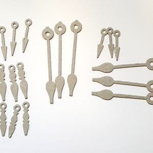 May include: A set of 14 clock hands made from cardboard. The hands are in various styles and sizes, including long and short hands, and hands with different shapes at the end. The hands are all a light brown colour.