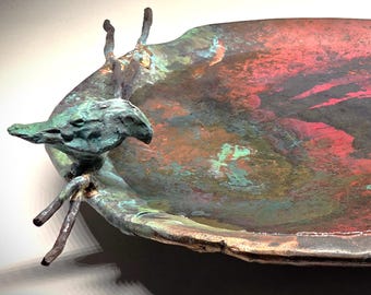 Puede incluir: Un cuenco de metal poco profundo y redondo con un borde irregular y texturizado. La superficie interior presenta una pátina vibrante y moteada de rojo intenso, marrón oscuro y verde azulado oxidado. Una figura de ave de metal en tono verdigris está unida al borde.