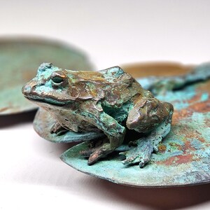 K&ouml;nnte beinhalten: Eine kleine gr&uuml;ne Froschskulptur, die auf einem kupferfarbenen Seerosenblatt sitzt. Der Frosch ist mit strukturierter Haut detailliert und hat ein realistisches Aussehen.